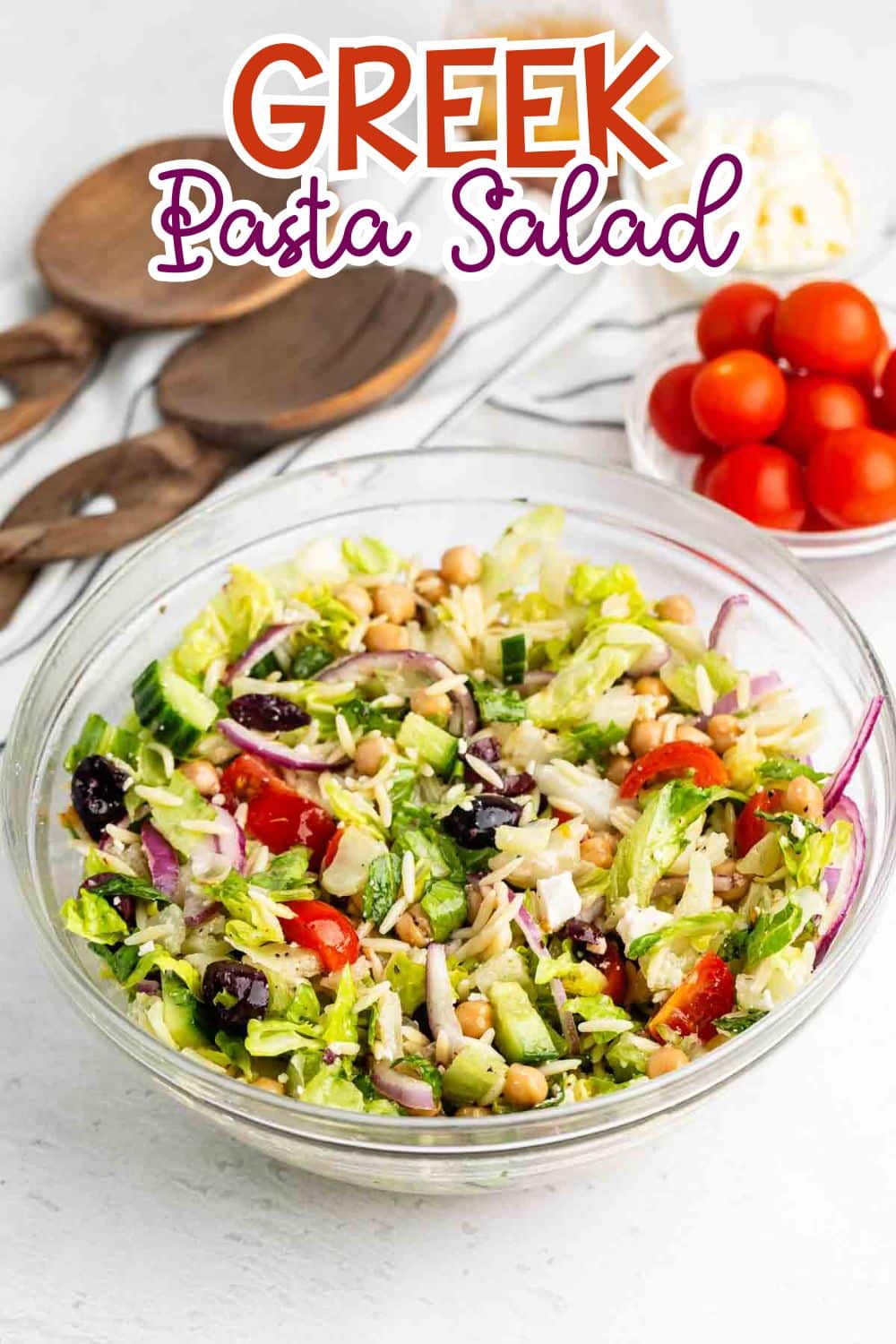 A glass bowl of Greek pasta salad with lettuce, cherry tomatoes, cucumbers, red onion, olives, chickpeas, and pasta. Wooden utensils and a bowl of cherry tomatoes are in the background. The text reads Greek Pasta Salad.