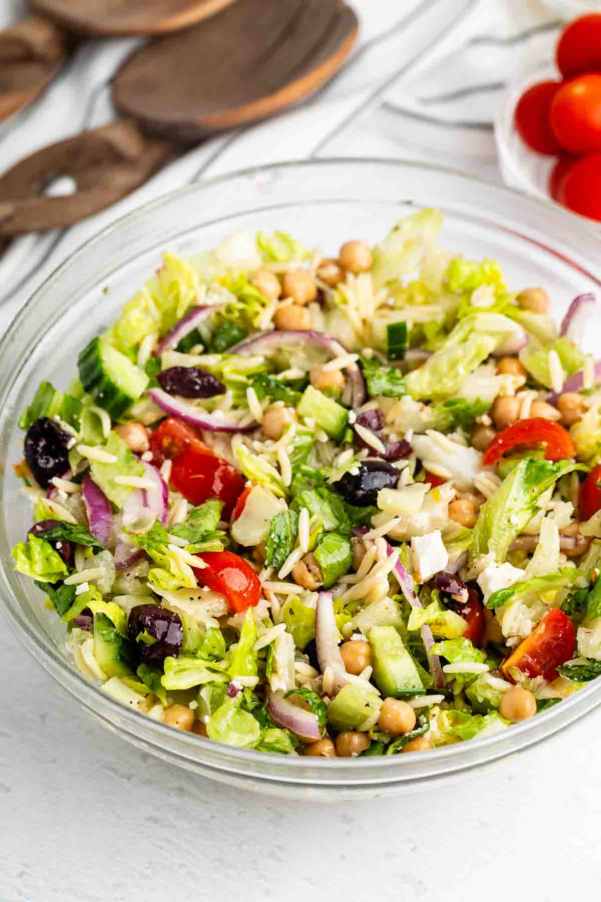 A glass bowl filled with Greek or Mediterranean salad containing lettuce, cherry tomatoes, cucumber, red onion, chickpeas, black olives, feta cheese, and orzo, with wooden utensils and tomatoes in the background.