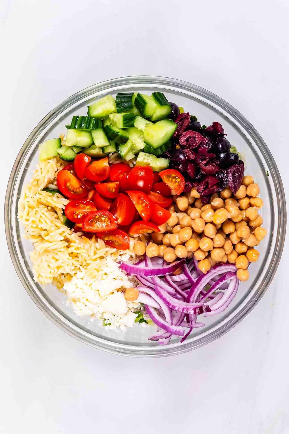 A glass bowl filled with chopped cucumber, cherry tomatoes, kalamata olives, chickpeas, sliced red onion, orzo pasta, and crumbled feta cheese, arranged in separate sections on a white background.