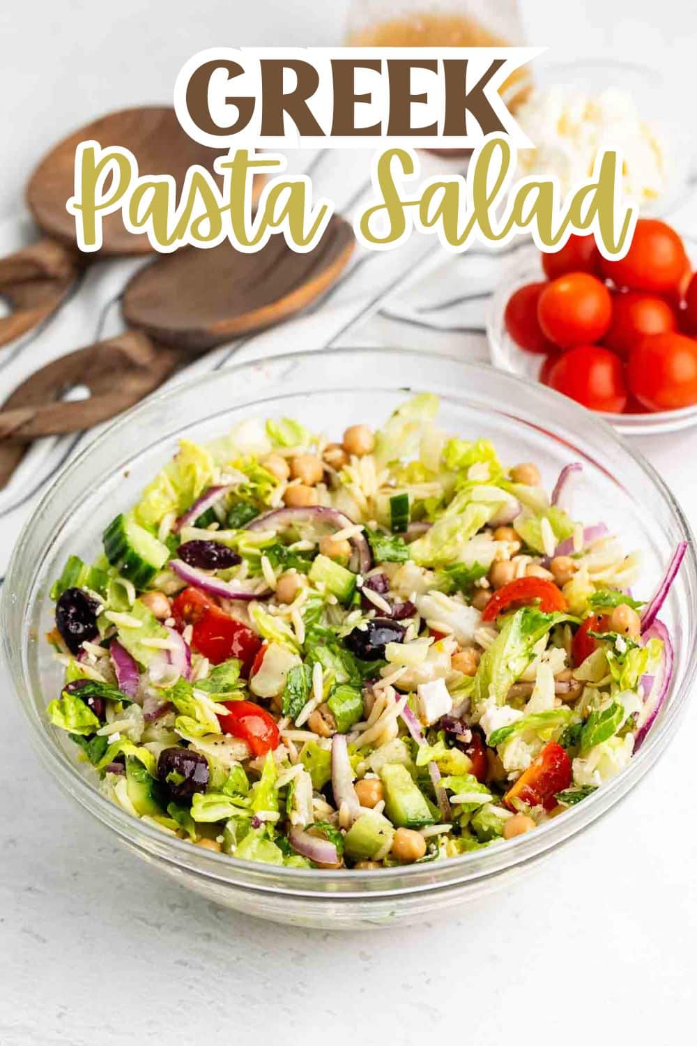 A glass bowl filled with Greek pasta salad containing lettuce, cherry tomatoes, cucumber, red onion, chickpeas, olives, and orzo. Wooden utensils and a bowl of cherry tomatoes are in the background. Text reads Greek Pasta Salad.
