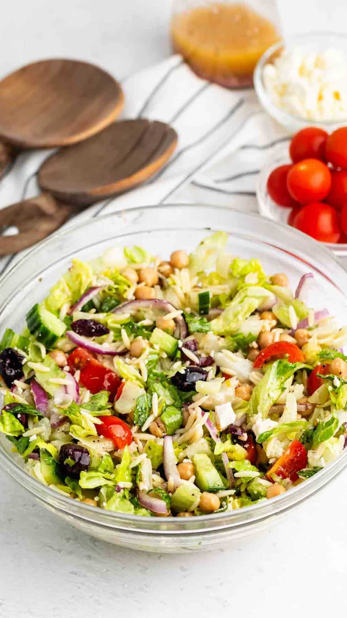 A glass bowl filled with a colorful salad containing lettuce, cherry tomatoes, cucumber, red onion, chickpeas, feta cheese, and olives. Wooden utensils, a bowl of cherry tomatoes, cheese, and dressing are in the background.