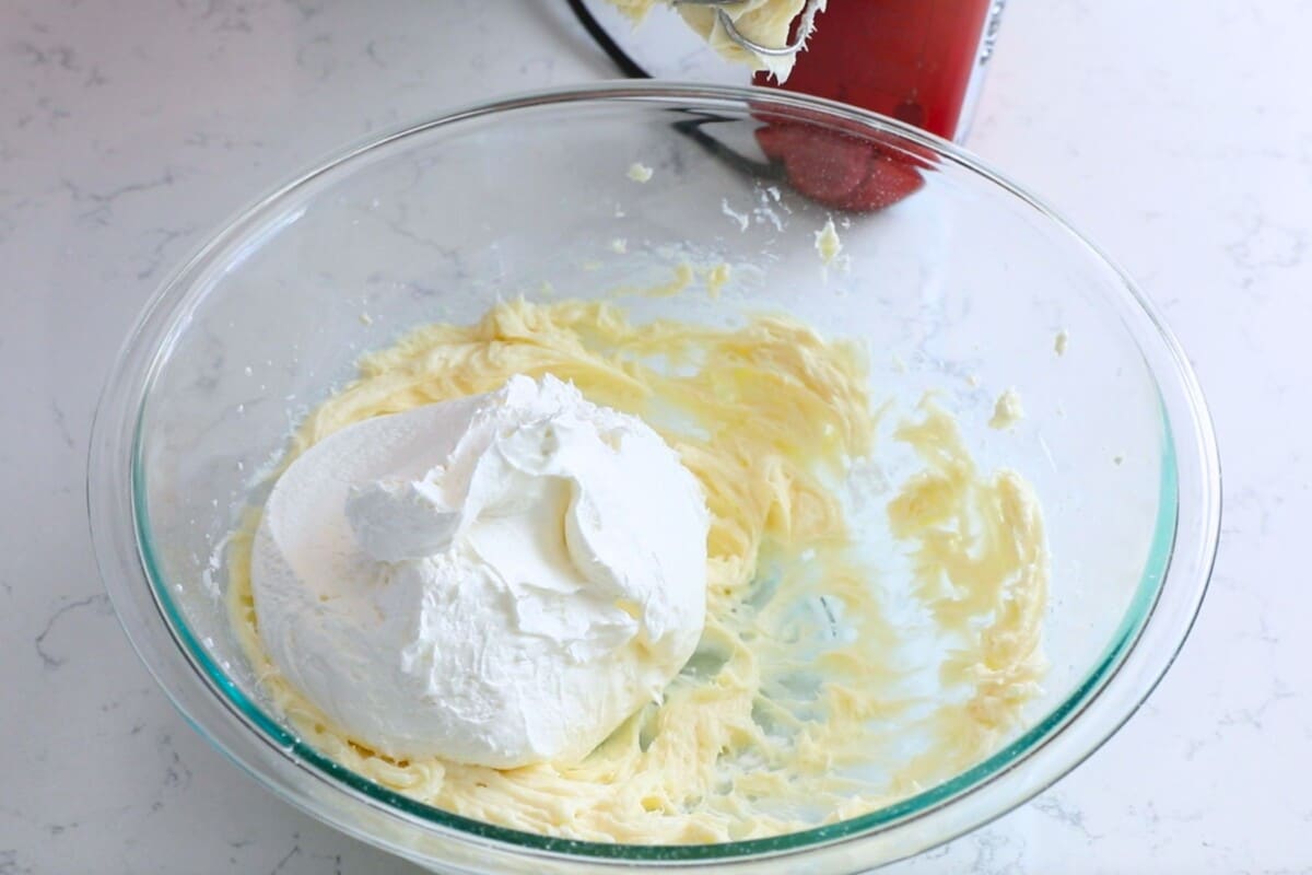 A glass mixing bowl with creamy yellow mixture and a large dollop of white whipped topping. An electric hand mixer with beaters is partially visible beside the bowl on a white countertop.