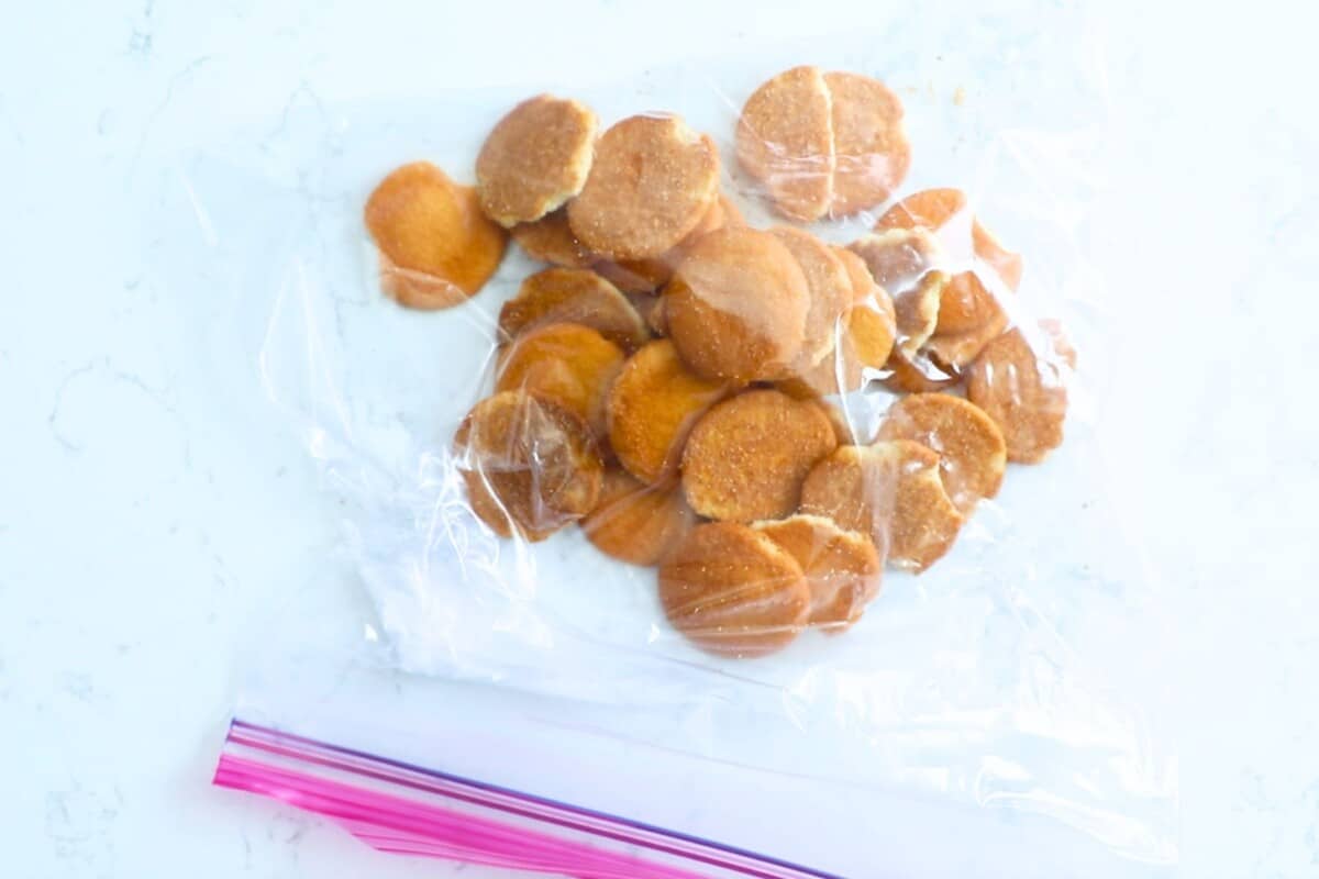Round vanilla wafers in a clear plastic resealable bag on a white surface; some of the cookies are whole, while others are broken or crumbled.