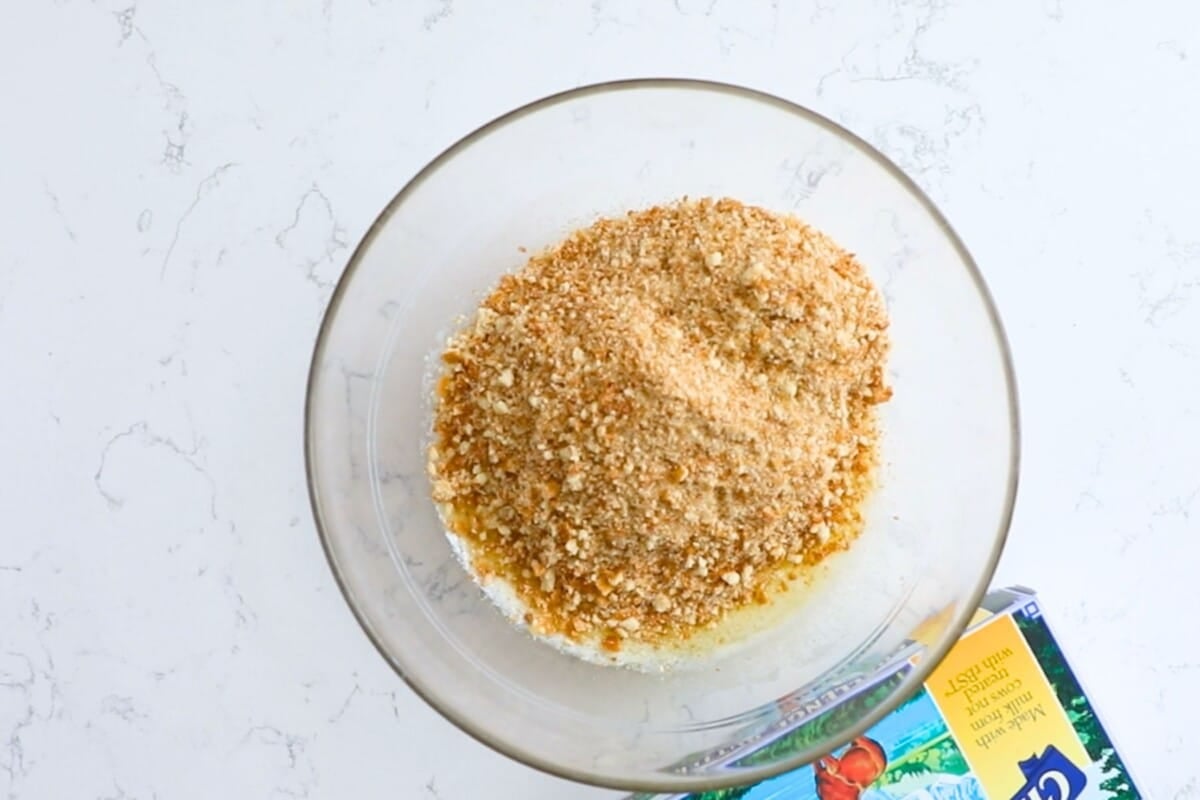 A glass bowl filled with breadcrumbs on top of melted butter sits on a white marble surface. Part of a butter box is visible in the lower right corner.