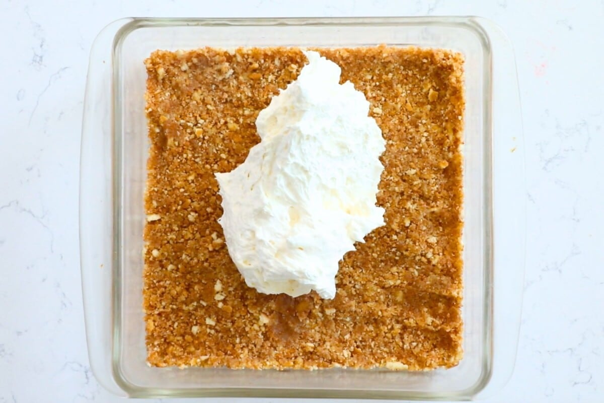 A glass baking dish with a graham cracker crust and a large dollop of whipped cream or cream cheese mixture in the center, viewed from above on a white marble surface.