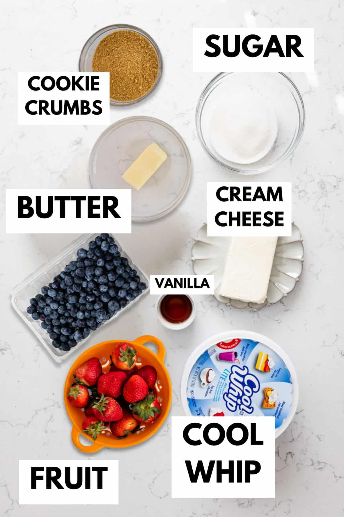 Top-down view of labeled ingredients on a white surface: cookie crumbs, sugar, butter, cream cheese, vanilla extract, Cool Whip, blueberries, and strawberries in bowls and containers.