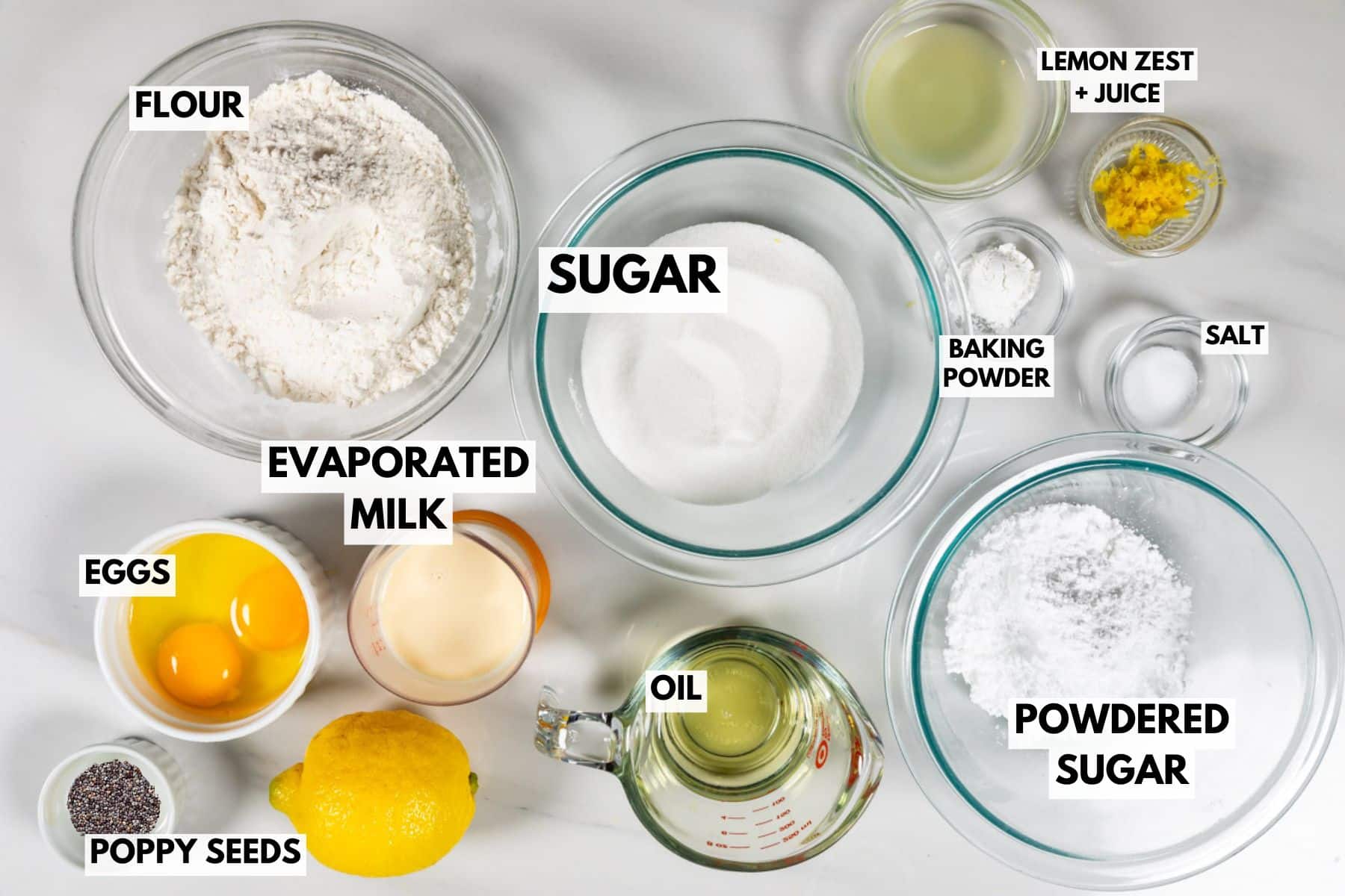 Top view of various baking ingredients in bowls on a white surface, including labeled flour, sugar, evaporated milk, eggs, poppy seeds, oil, powdered sugar, lemon zest and juice, baking powder, salt, and a whole lemon.