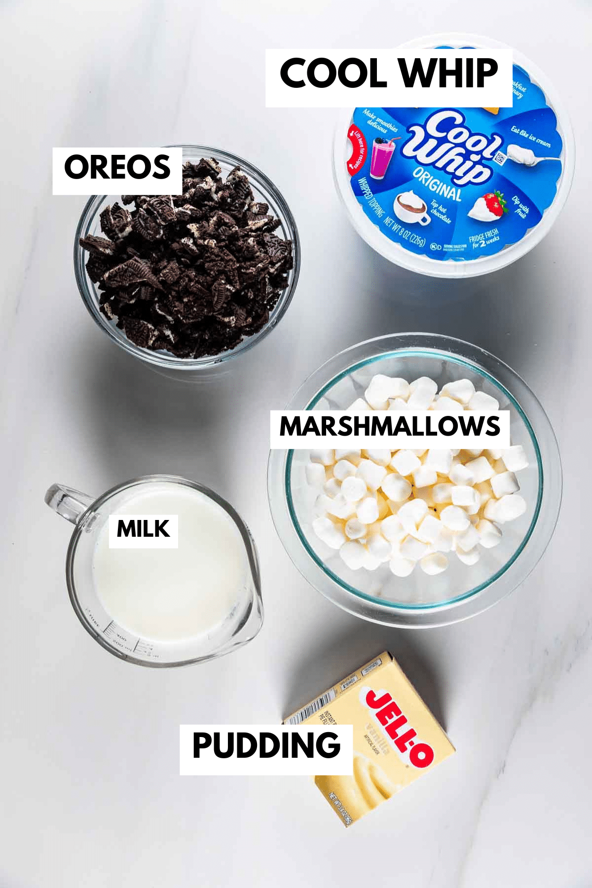Overhead view of five ingredients labeled: a tub of Cool Whip, a bowl of crushed Oreos, a bowl of marshmallows, a glass measuring cup of milk, and a box of Jello pudding.