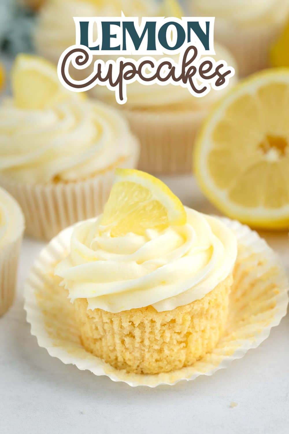 A lemon cupcake with swirled frosting and a lemon wedge on top sits in a cupcake liner. Other cupcakes and a sliced lemon are in the background. Text at the top reads Lemon Cupcakes.