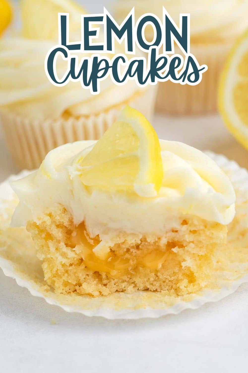 A lemon cupcake with creamy frosting and a lemon slice on top, partially unwrapped with a bite taken out, revealing a lemon filling. Other cupcakes and lemon slices are blurred in the background. Text reads “Lemon Cupcakes.”.