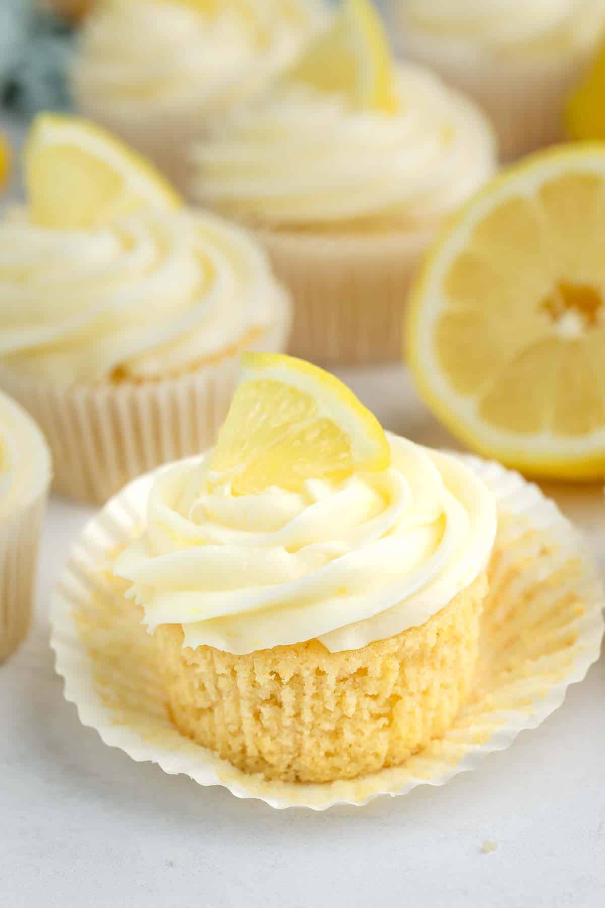 A lemon cupcake with creamy white frosting and a small lemon wedge on top, sitting on an unwrapped cupcake liner. Other cupcakes and a halved lemon are visible in the background.