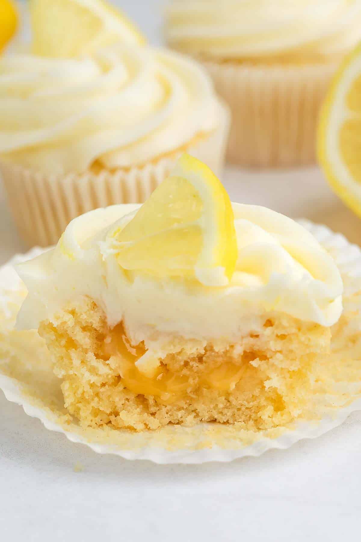 A lemon cupcake with creamy frosting and a lemon slice on top sits partially unwrapped, showing a gooey lemon filling inside. Other frosted cupcakes and a lemon wedge are in the background.