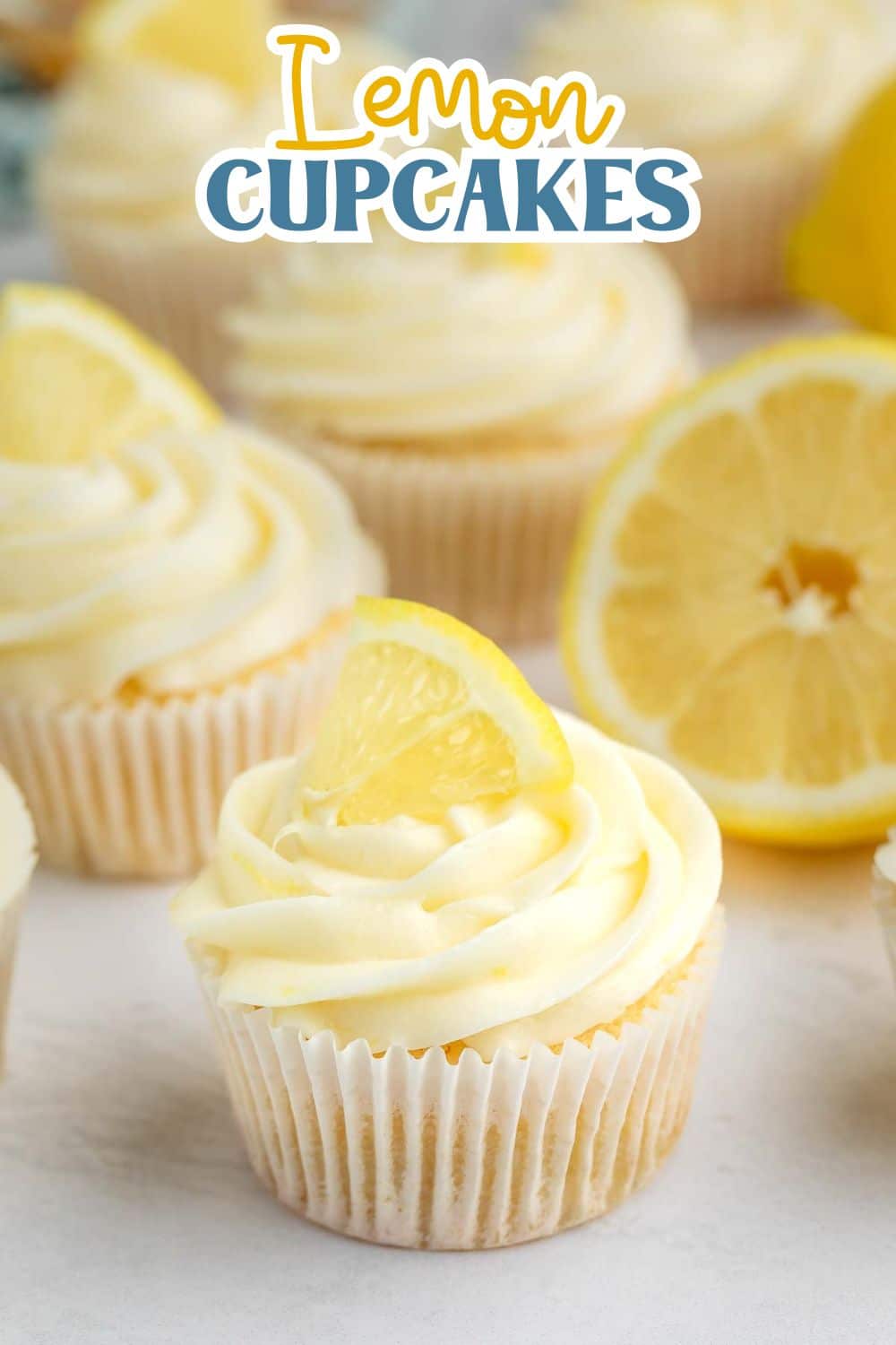 Cupcakes with creamy white frosting, each topped with a lemon slice, sit on a light surface. In the background, there is a sliced lemon and the text “Lemon Cupcakes” at the top.