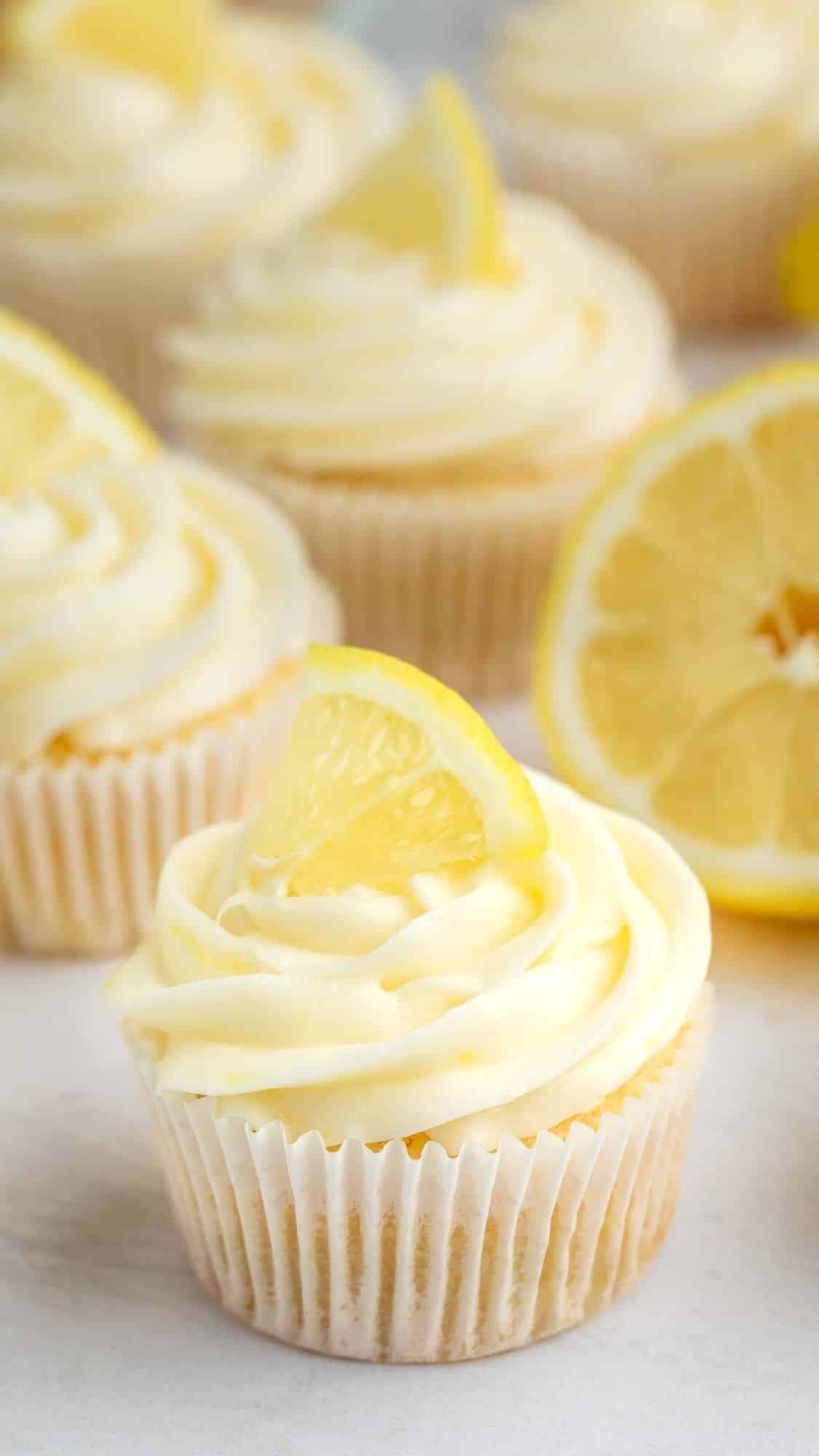 A lemon cupcake with creamy white frosting topped with a slice of lemon, surrounded by more cupcakes and a halved lemon in the background.