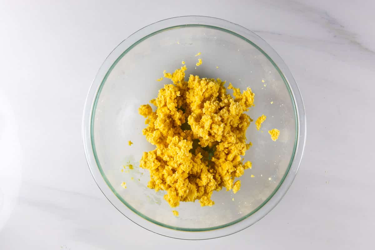 A clear glass bowl containing a mixture of crumbled yellow dough sits on a white marble surface.