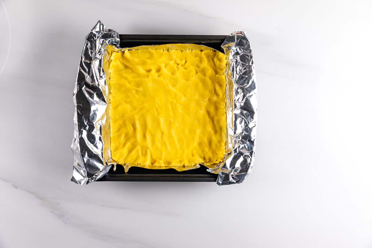 A baking pan lined with aluminum foil holds an unbaked, evenly spread yellow dough or crust on a white marble surface.