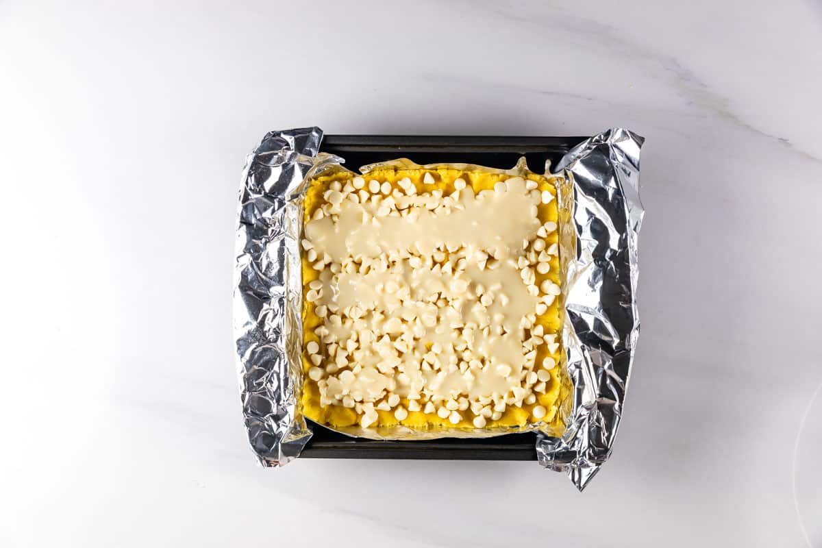 A square baking pan lined with foil holds an unbaked dessert topped with white chocolate chips and a creamy layer, placed on a white marble surface.