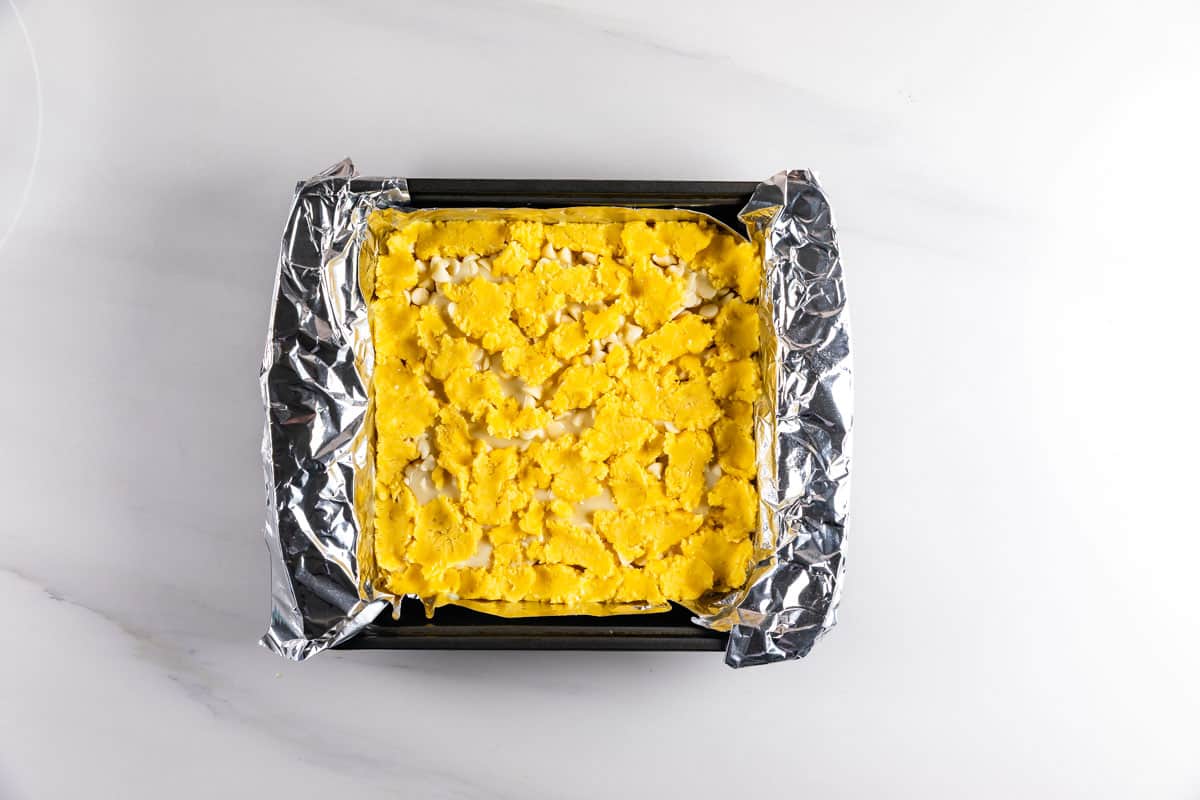 A square baking pan lined with aluminum foil, filled with an unbaked mixture that appears to be a yellow crumbly dough, sitting on a white marble surface.