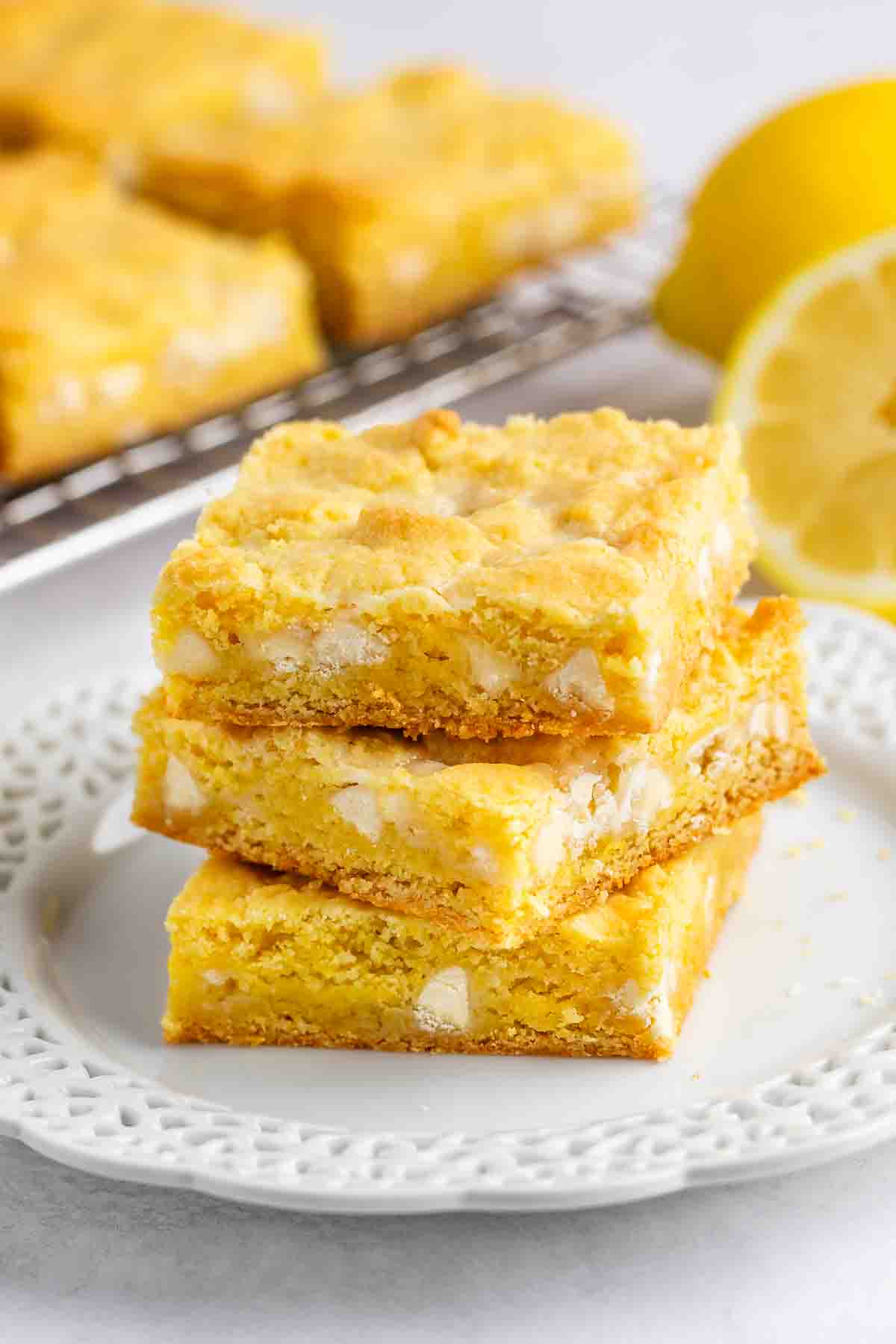 Three lemon blondie bars stacked on a white decorative plate, with a sliced lemon and more blondies on a cooling rack in the background. The bars have a golden, crumbly texture and visible white chocolate chunks.