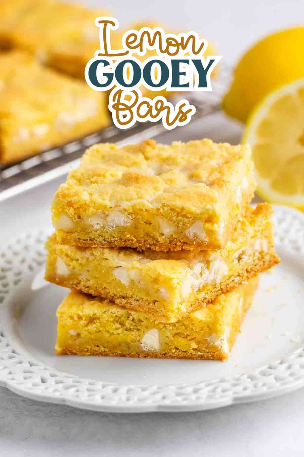 Three lemon gooey bars are stacked on a white plate, with a sliced lemon and a baking rack of bars in the background. The text Lemon Gooey Bars is displayed above the stack.