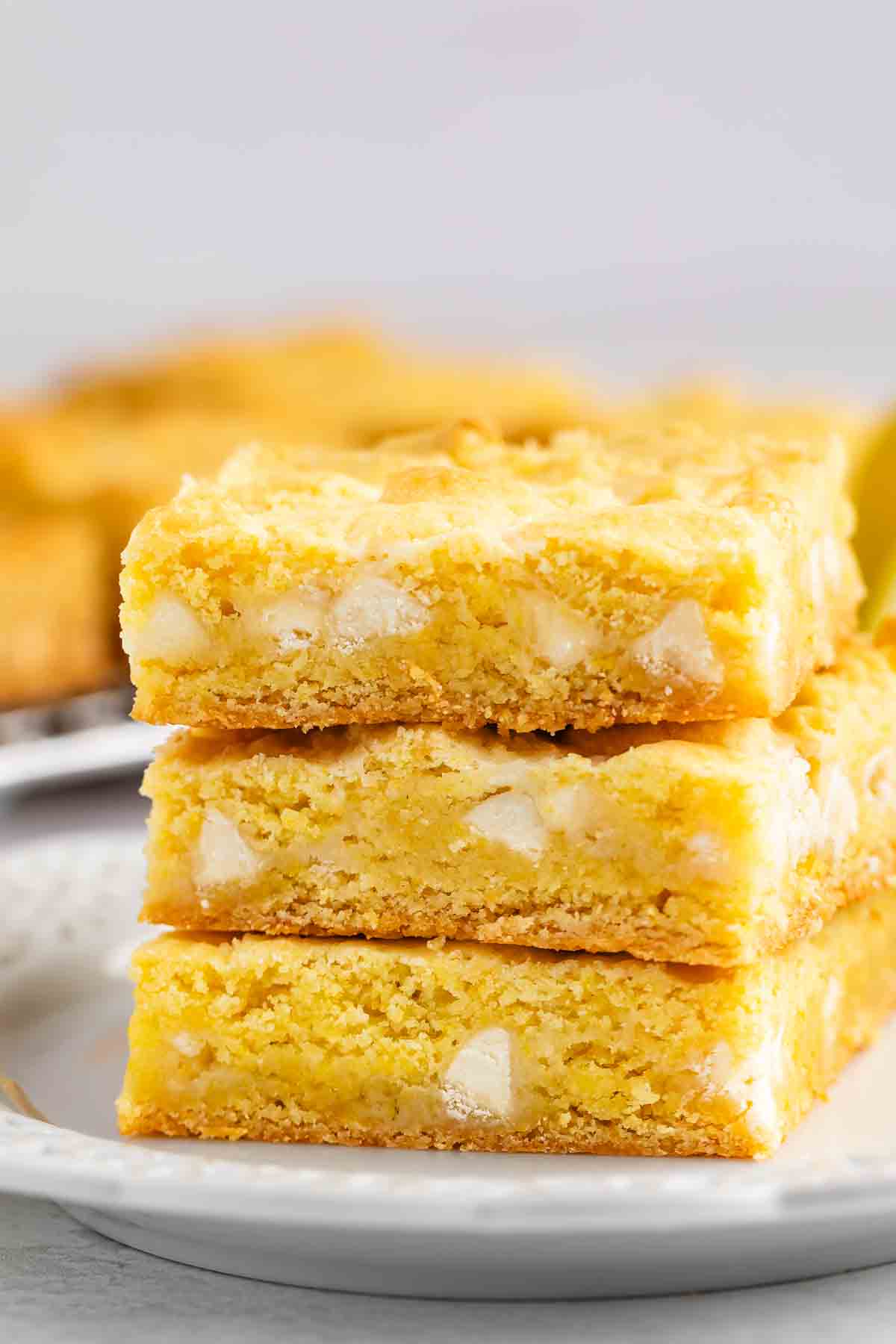 Three golden, chewy blondie bars with visible white chocolate chunks are stacked on a white plate, showcasing their soft texture and slightly crumbly edges.