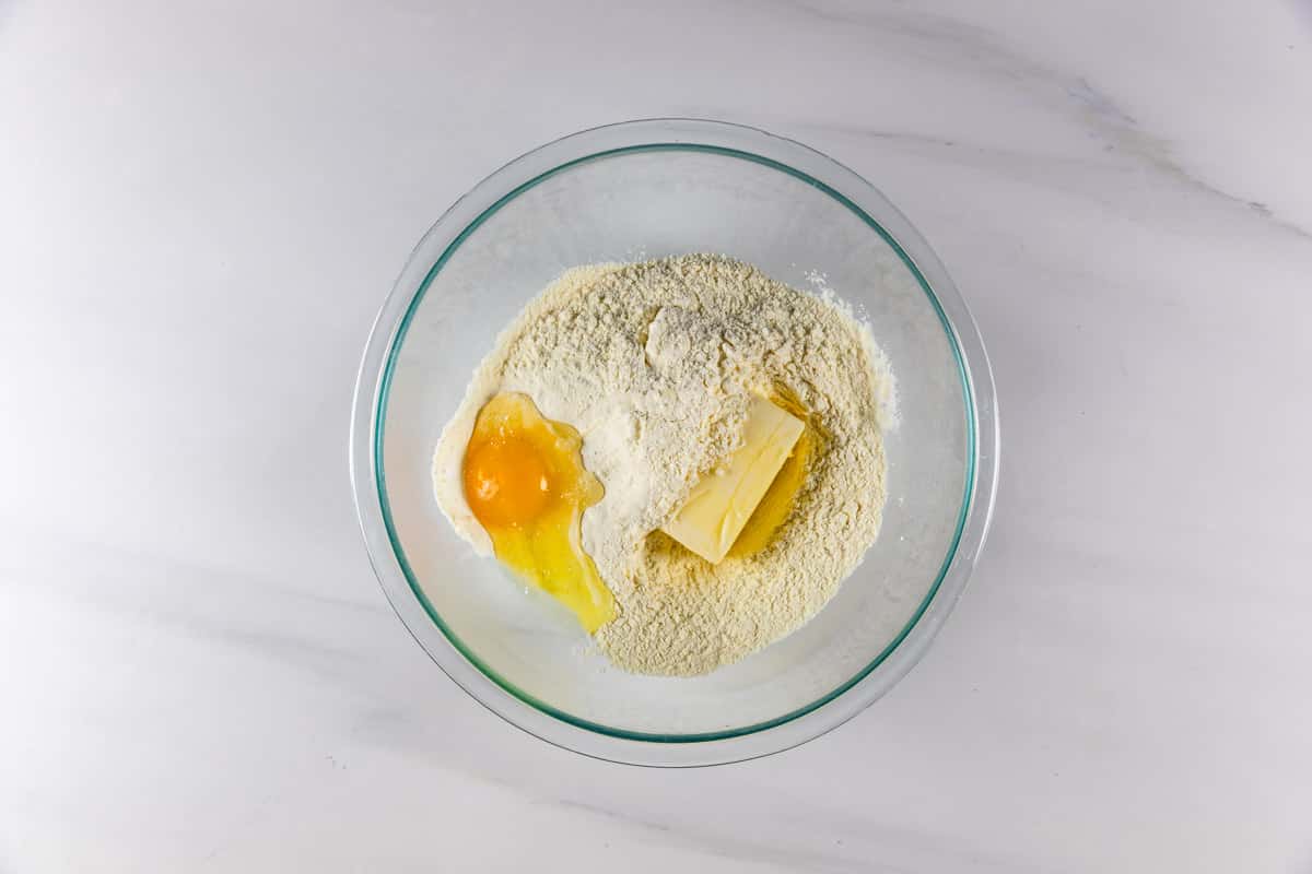 A glass bowl containing flour, a cracked egg, and a piece of butter sits on a white countertop, ready for mixing.