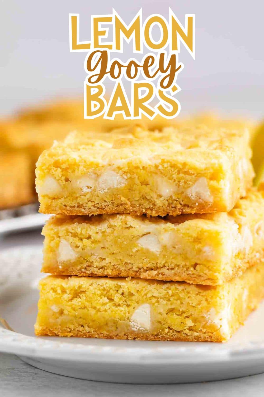 A stack of three lemon gooey bars with visible white chocolate chunks sits on a white plate. In the background, more bars are slightly blurred. Bold text above reads Lemon Gooey Bars.