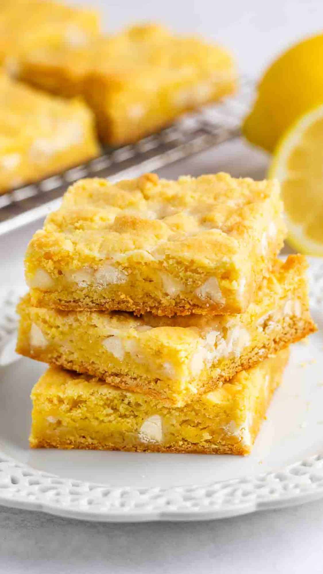Three square lemon bars stacked on a white plate, with a bright yellow color and visible chunks, set in front of a wire rack and a sliced lemon in the background.