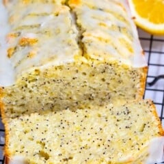 A loaf of lemon poppy seed bread with a white glaze sits on a cooling rack. One slice is cut and slightly pulled out. A half lemon is placed beside the loaf. The bread has visible poppy seeds throughout.