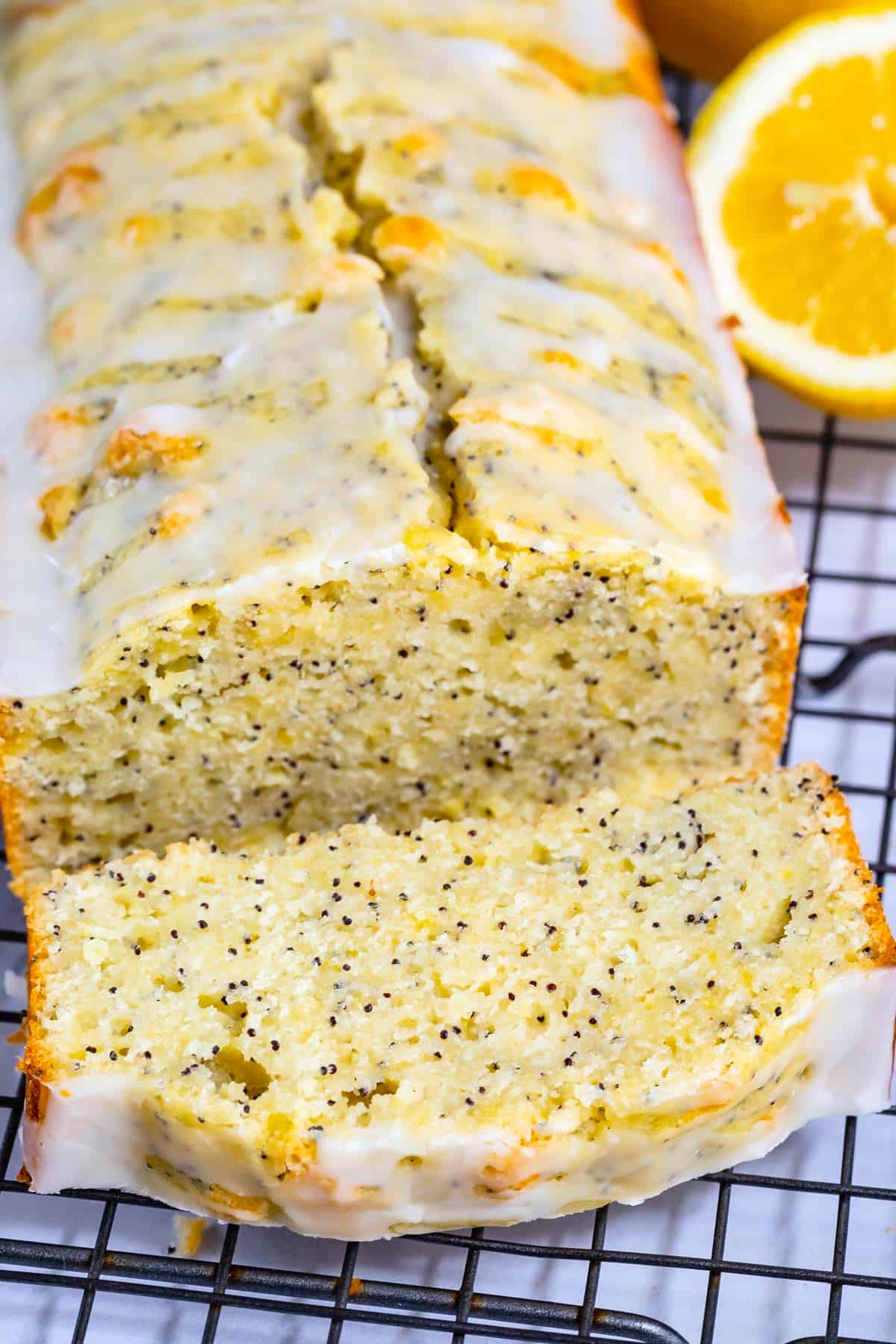 A loaf of lemon poppy seed bread with a white glaze sits on a cooling rack. One slice is cut and slightly pulled out. A half lemon is placed beside the loaf. The bread has visible poppy seeds throughout.