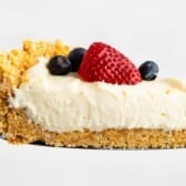 A slice of creamy no bake cheesecake with a crumbly graham cracker crust, topped with a halved strawberry and two blueberries, held up on a metal spatula against a white background.
