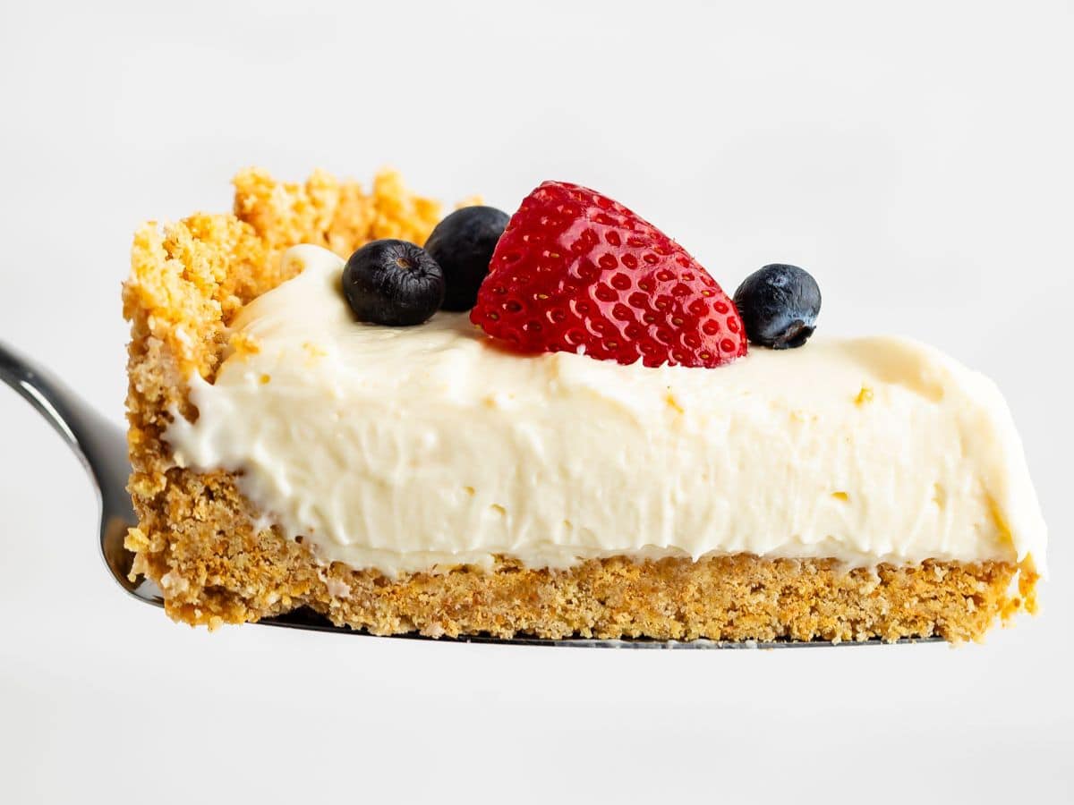A slice of creamy no bake cheesecake with a crumbly graham cracker crust, topped with a halved strawberry and two blueberries, held up on a metal spatula against a white background.