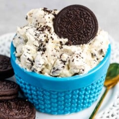 A blue bowl filled with cookies and cream dip, topped with a whole chocolate sandwich cookie, sits on a white plate with additional cookies and a gold spoon beside it.