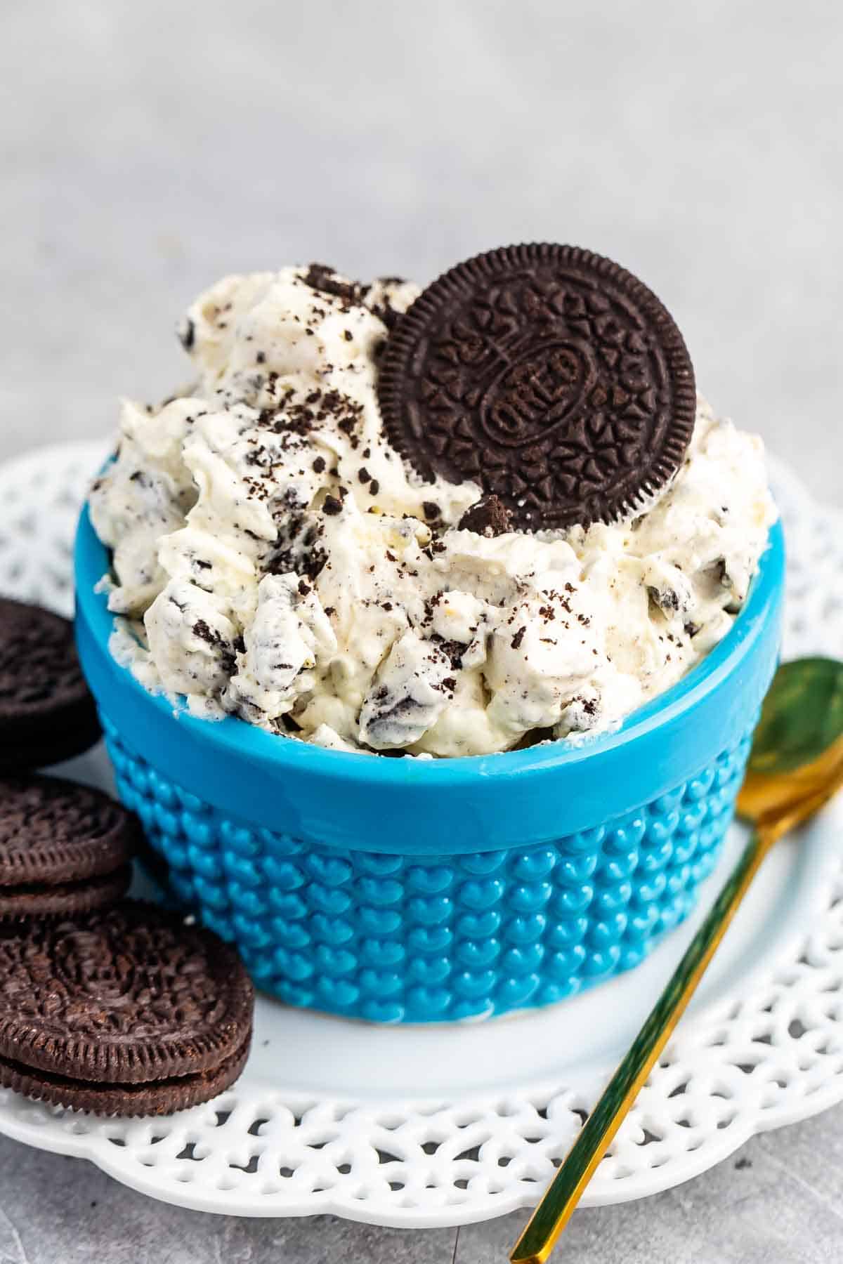 A blue bowl filled with cookies and cream dip, topped with a whole chocolate sandwich cookie, sits on a white plate with additional cookies and a gold spoon beside it.