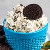 A blue textured bowl filled with creamy Oreo dessert, topped with crumbled Oreos and a whole Oreo cookie. A few Oreos and a gold spoon are placed beside the bowl on a white plate.