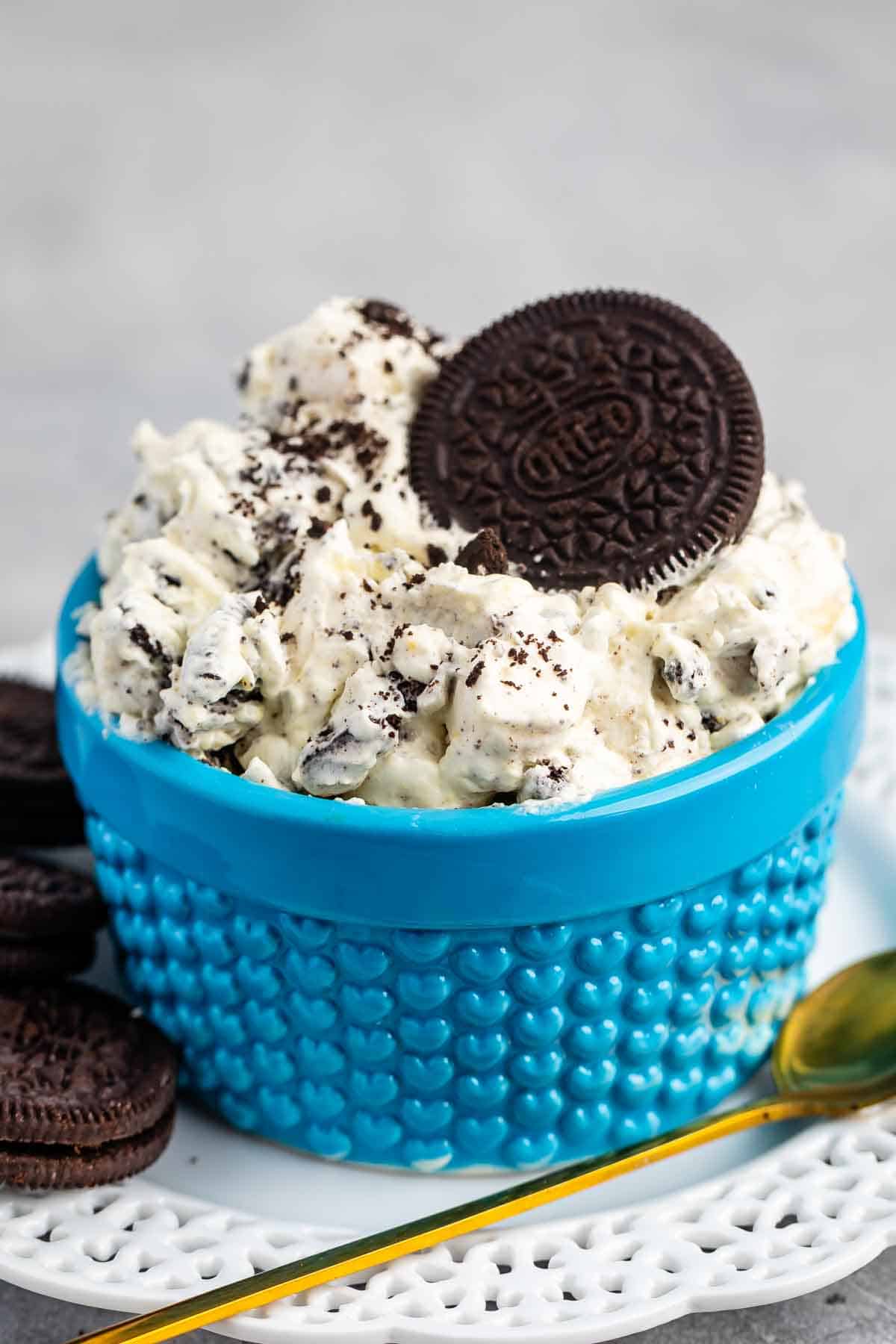 A blue textured bowl filled with creamy Oreo dessert, topped with crumbled Oreos and a whole Oreo cookie. A few Oreos and a gold spoon are placed beside the bowl on a white plate.