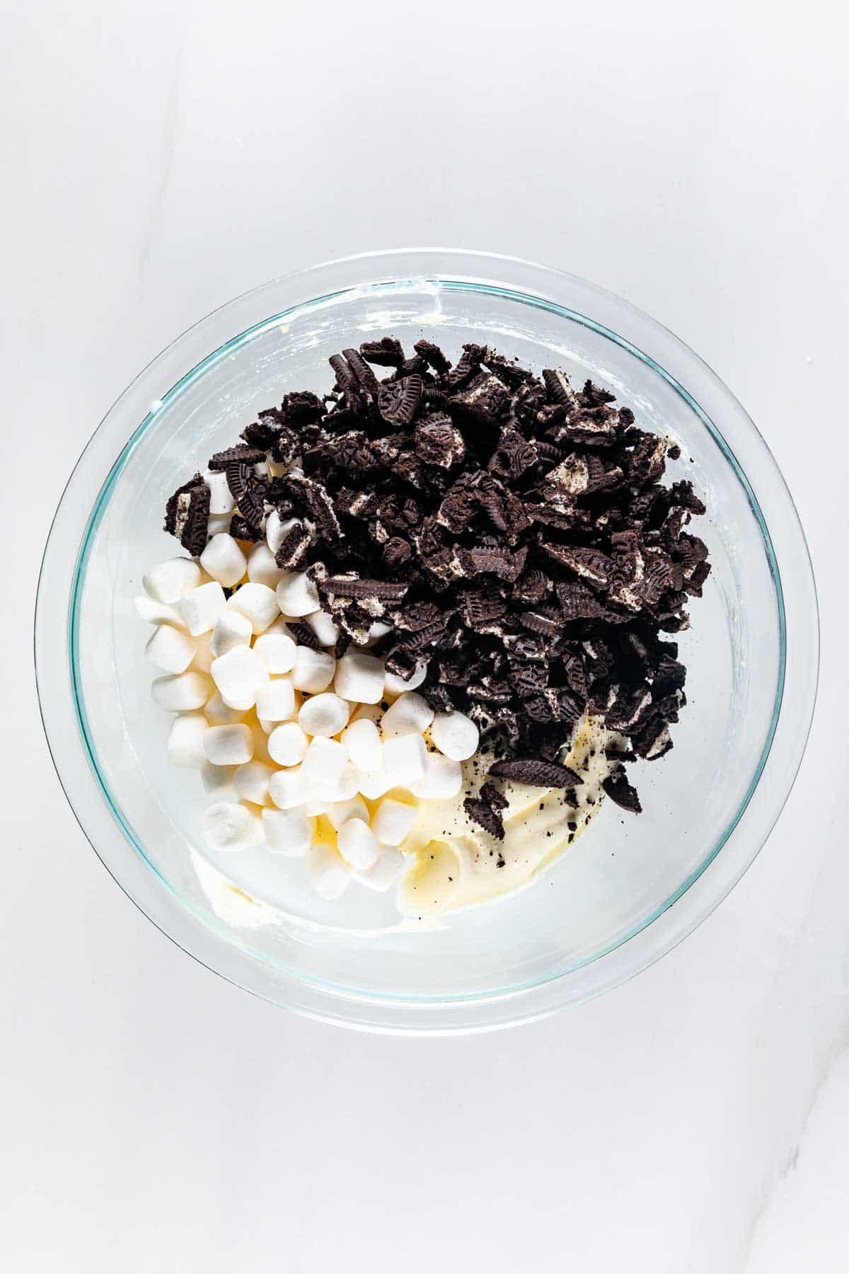 A glass bowl containing mini marshmallows, chopped chocolate sandwich cookies, and a dollop of cream cheese on a white surface.
