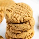 A stack of five peanut butter cookies with a crisscross fork pattern on top, placed on a white surface with more cookies in the background.