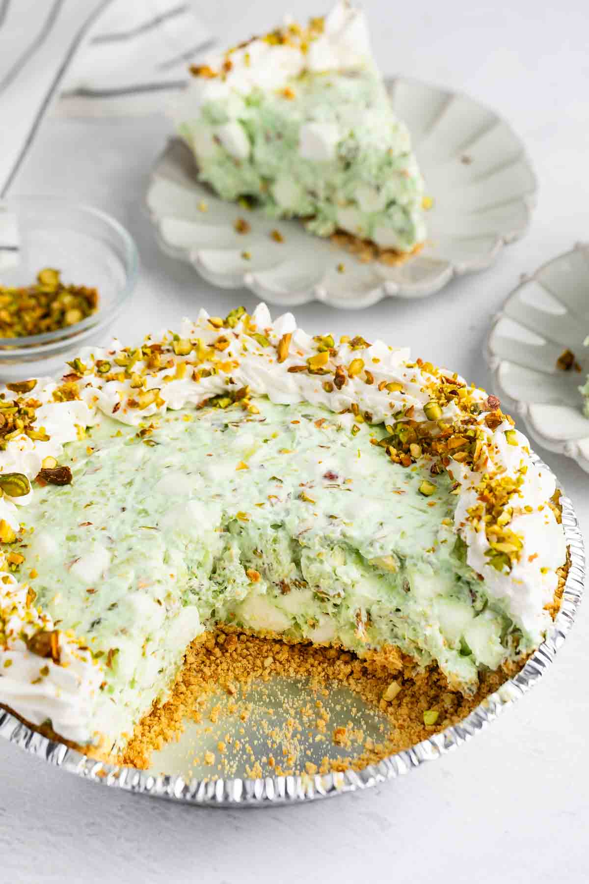 A partially sliced pistachio pie with a graham cracker crust, creamy green filling, whipped topping, and chopped pistachios, served on a white plate with a piece taken out and another slice on a plate in the background.
