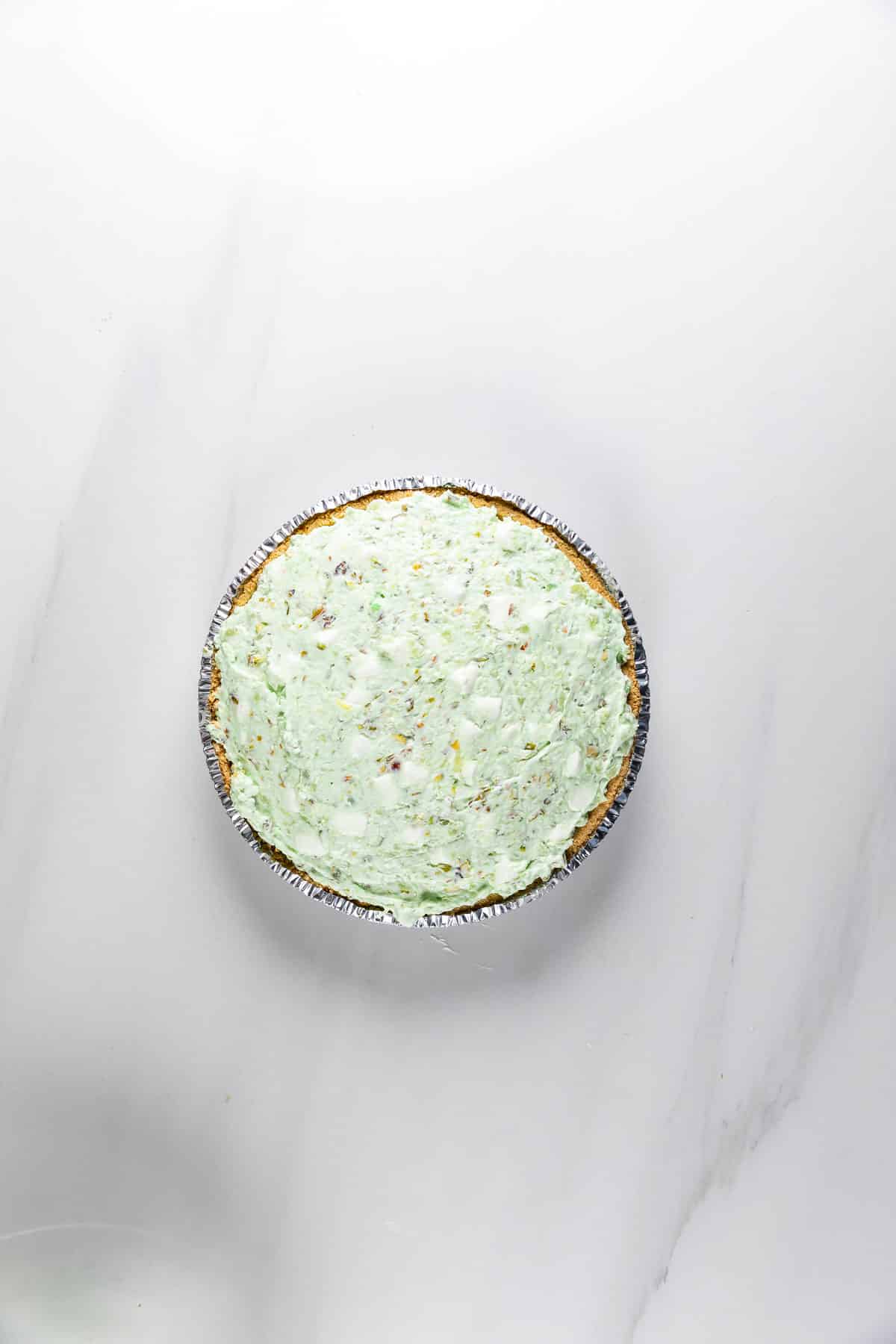 A round pie with a light green creamy filling and small specks, set in a graham cracker crust and placed on a white marble surface.