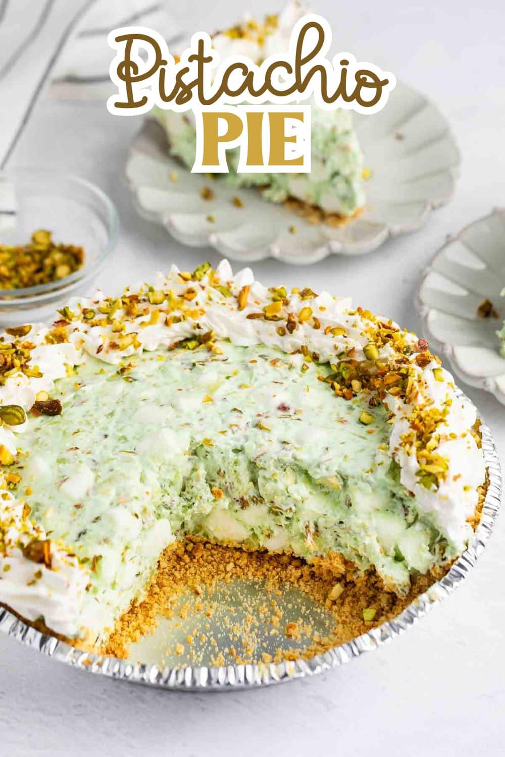 A pistachio pie with a graham cracker crust, creamy green filling, and whipped topping, garnished with chopped pistachios. A slice is missing from the pie, and a plated slice is in the background. Text reads Pistachio Pie.