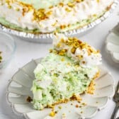 A slice of creamy green pistachio pie topped with whipped cream and chopped pistachios sits on a white plate, with the rest of the pie and utensils visible in the background.