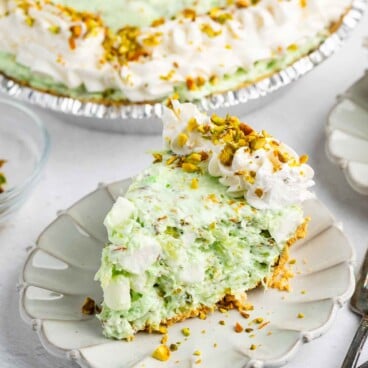 A slice of creamy green pistachio pie topped with whipped cream and chopped pistachios sits on a white plate, with the rest of the pie and utensils visible in the background.
