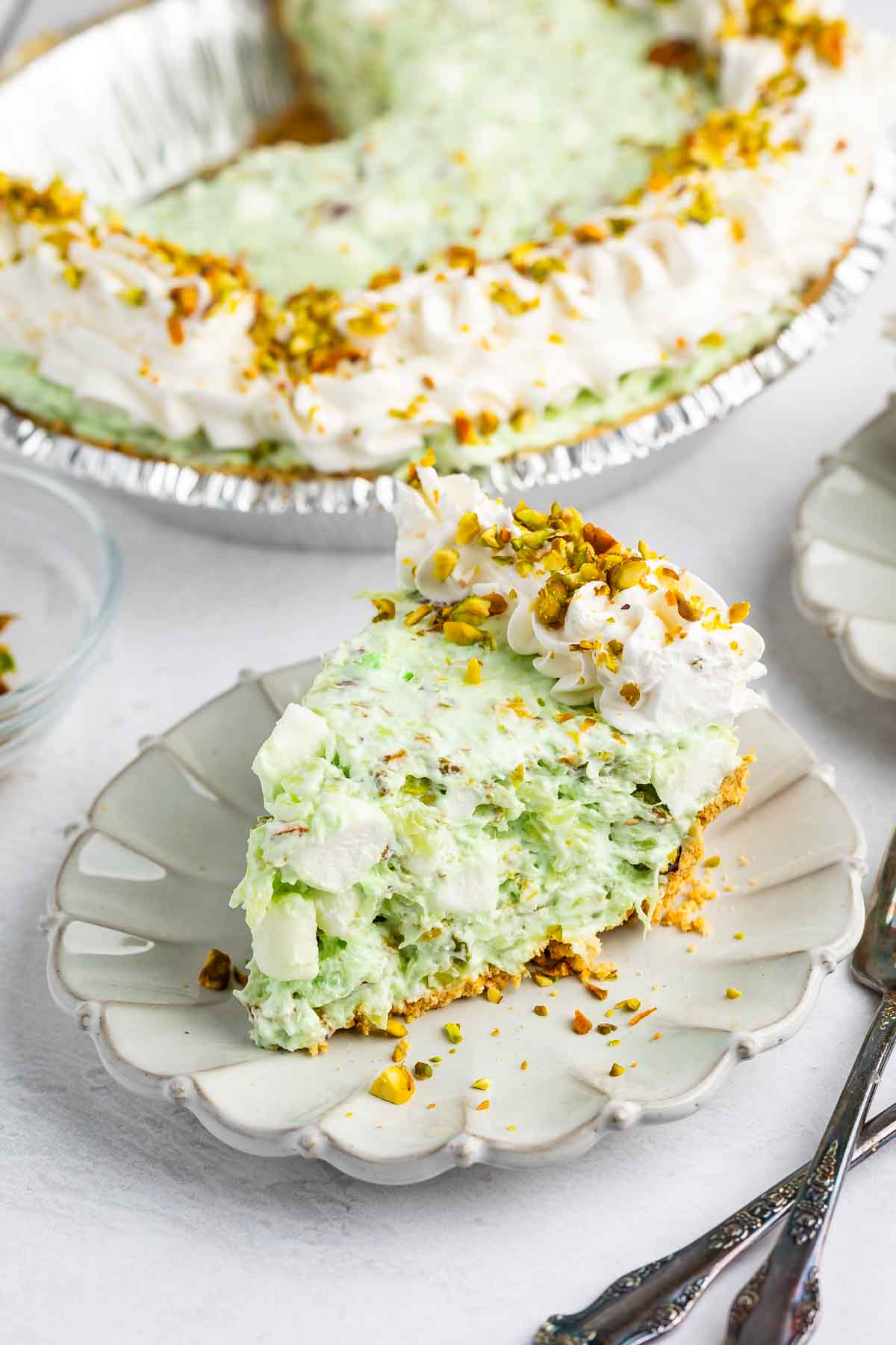 A slice of creamy green pistachio pie topped with whipped cream and chopped pistachios sits on a white plate, with the rest of the pie and utensils visible in the background.