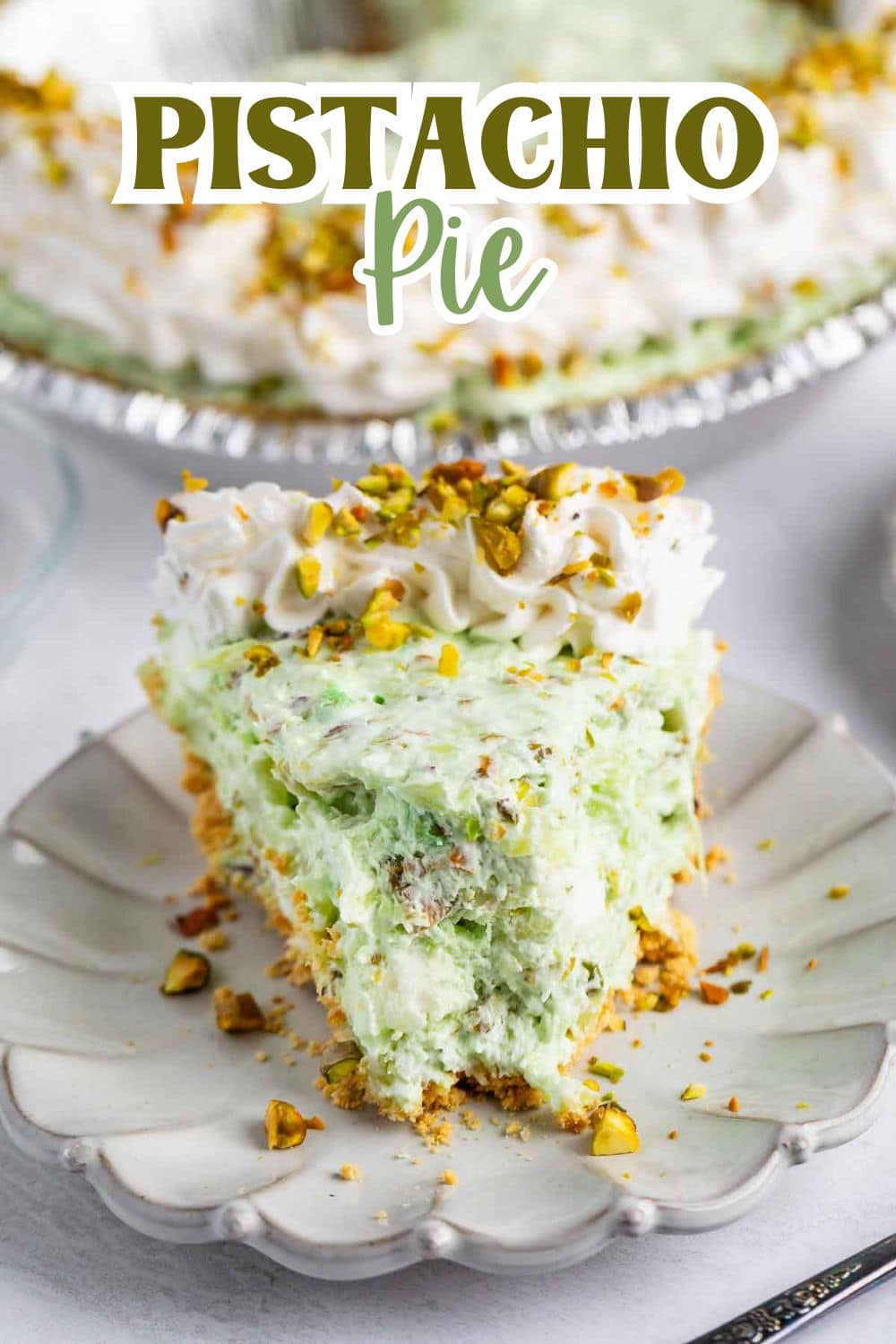 A slice of creamy pistachio pie topped with whipped cream and chopped pistachios sits on a decorative plate, with the rest of the pie in the background. Text above reads Pistachio Pie.