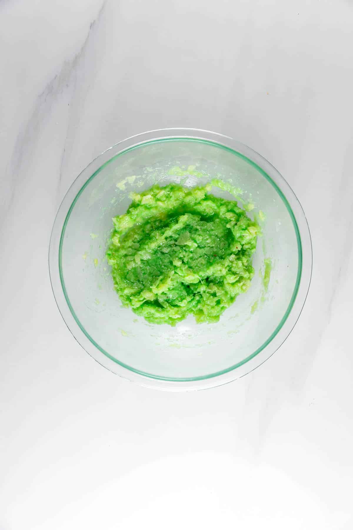 A clear glass bowl filled with bright green mashed avocado sits on a white marble surface.