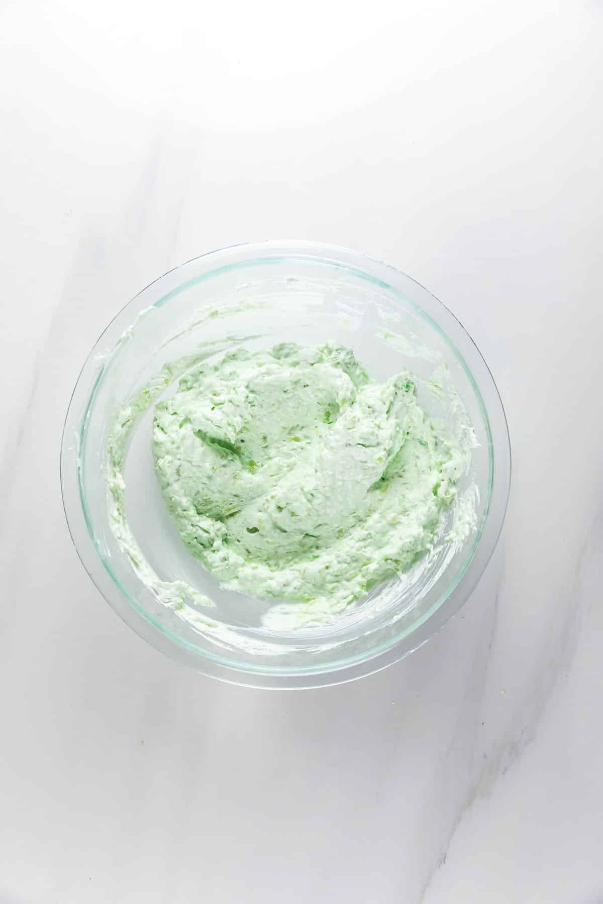 A glass bowl filled with a light green, creamy mixture sits on a white marble surface.