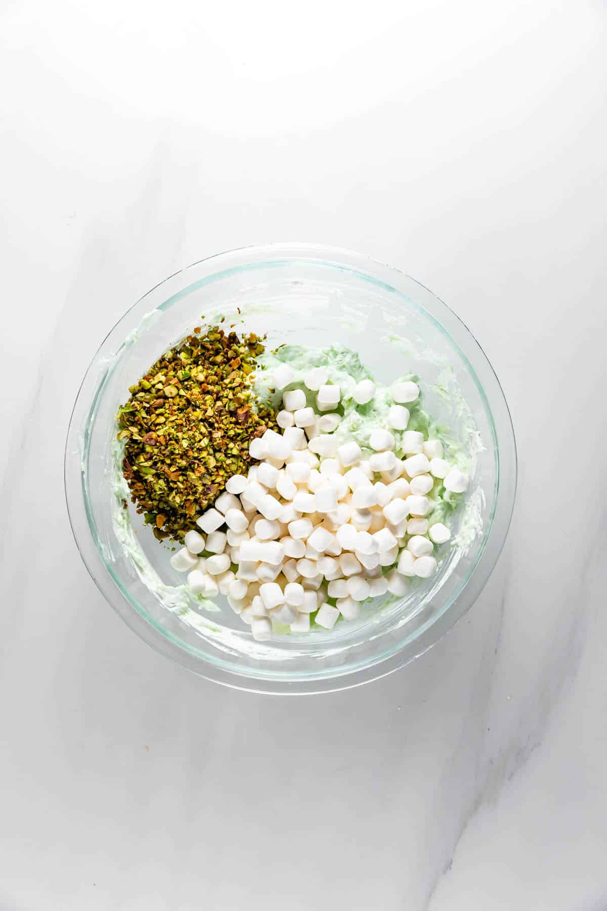 A glass bowl on a white surface contains a mixture of chopped pistachios, mini marshmallows, and a pale green creamy base, ready to be mixed together.