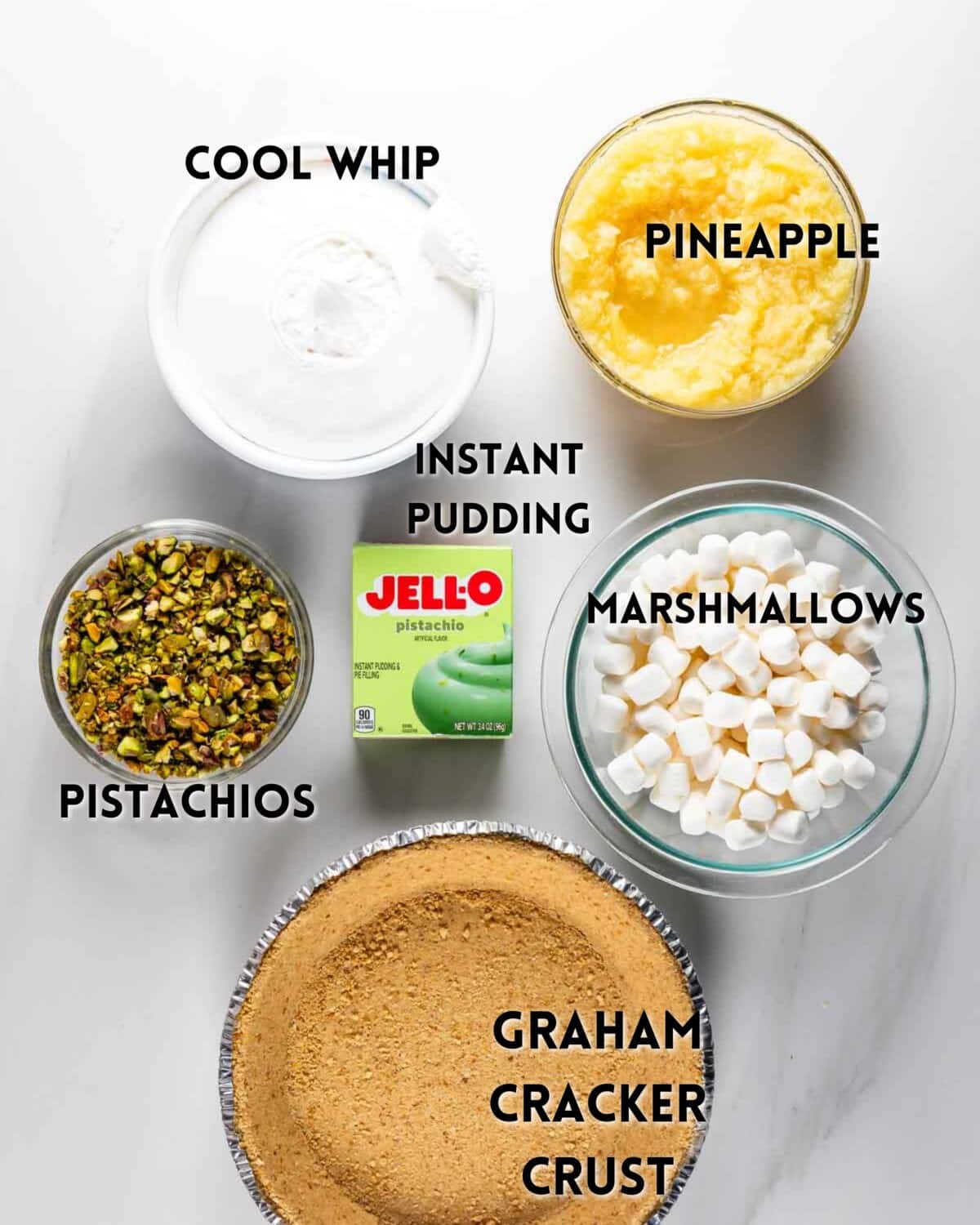 A top-down view of ingredients for a dessert: Cool Whip, crushed pineapple, instant pistachio pudding mix, mini marshmallows, chopped pistachios, and a graham cracker crust, each labeled.