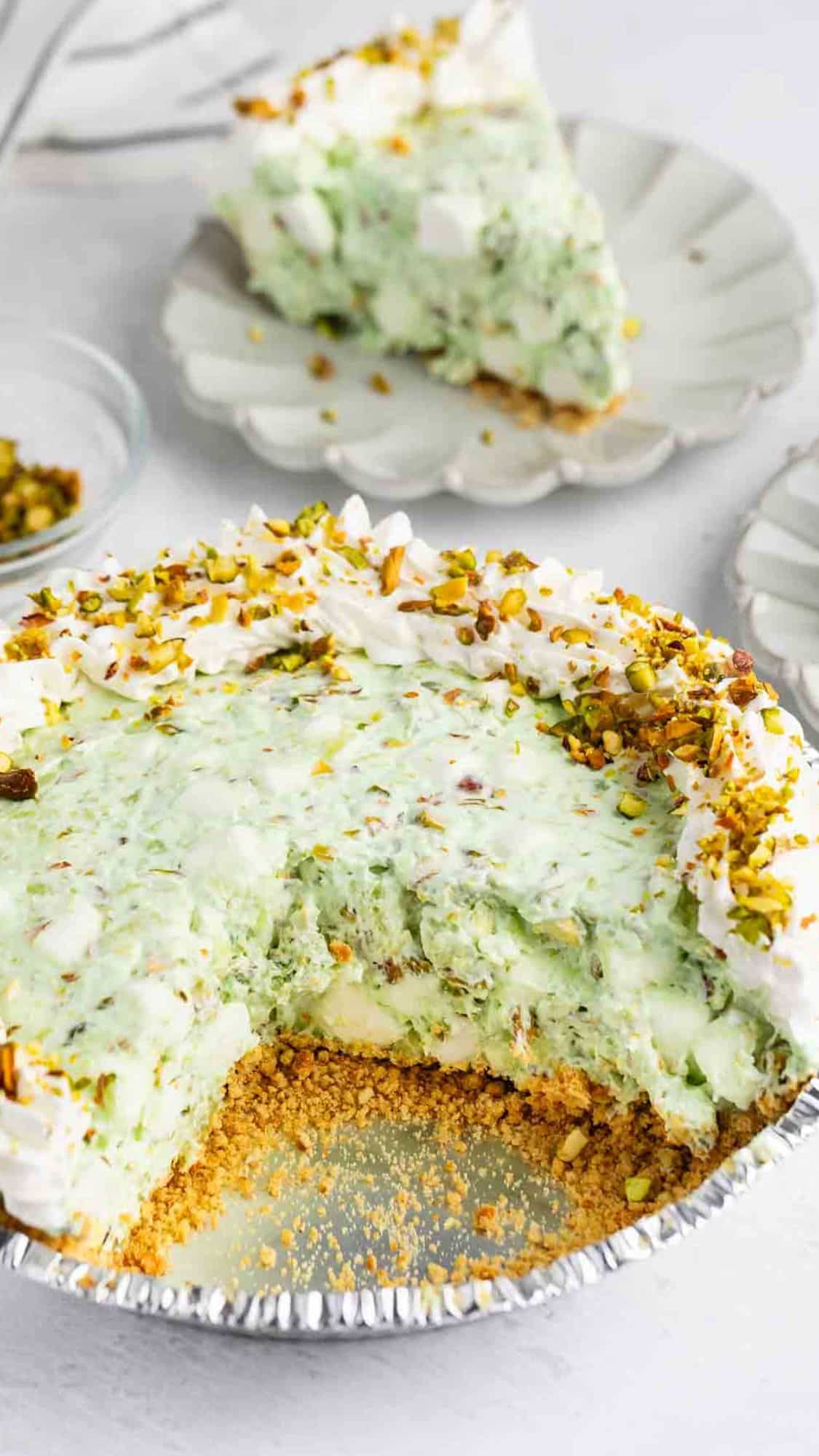 A partially sliced pistachio pie with a graham cracker crust, creamy green filling, whipped topping, and chopped pistachios on top. A slice of the pie is served on a plate in the background.
